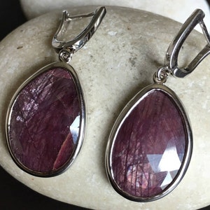 May include: A pair of silver dangle earrings with a teardrop-shaped purple gemstone. The earrings have a silver hook closure.