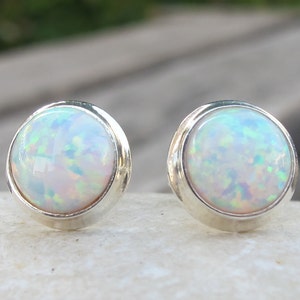 May include: A pair of round opal stud earrings. Each earring features a white opal stone with iridescent flashes of color, set in a silver bezel. The earrings are displayed on a white surface, with a blurred background.