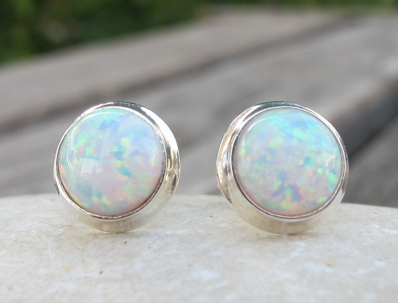 May include: A pair of round opal stud earrings. Each earring features a white opal stone with iridescent flashes of color, set in a silver bezel. The earrings are displayed on a white surface, with a blurred background.