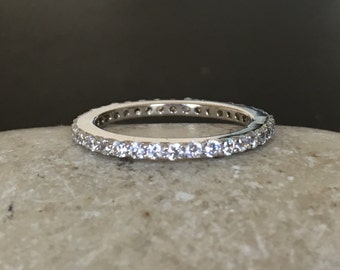 Puede incluir: Un anillo de banda de plata con una fila de pequeñas piedras de circonita cúbica transparentes engastadas alrededor de toda la banda.