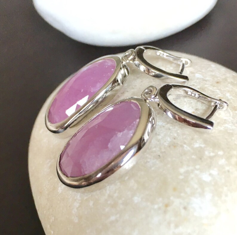 K&ouml;nnte beinhalten: Ein Paar silberne Ohrringe mit rosafarbenen, ovalen Edelsteinen. Die Ohrringe haben ein schlichtes Design mit einem Hakenverschluss.
