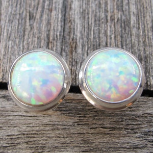 May include: A pair of round stud earrings featuring iridescent opal stones set in silver. The opals display a play of color with flashes of green, blue, and pink. The earrings are displayed on a weathered wooden surface.