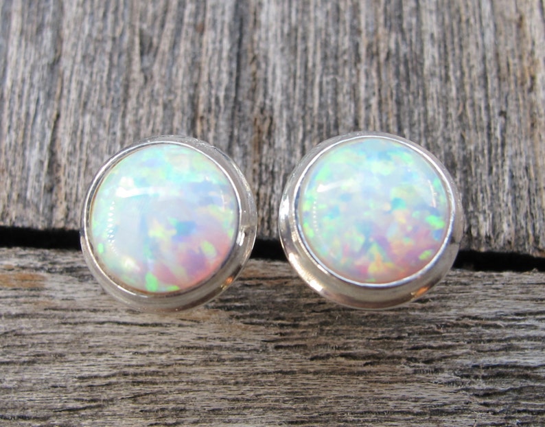 May include: A pair of round stud earrings featuring iridescent opal stones set in silver. The opals display a play of color with flashes of green, blue, and pink. The earrings are displayed on a weathered wooden surface.