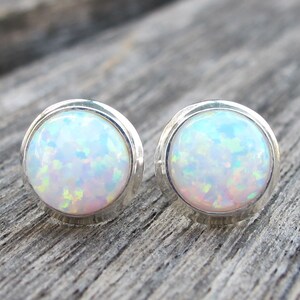 May include: A pair of round stud earrings featuring iridescent opal stones set in silver bezels. The opals display a mix of colors, including white, blue, and hints of pink and green. The earrings are displayed on a wooden surface.