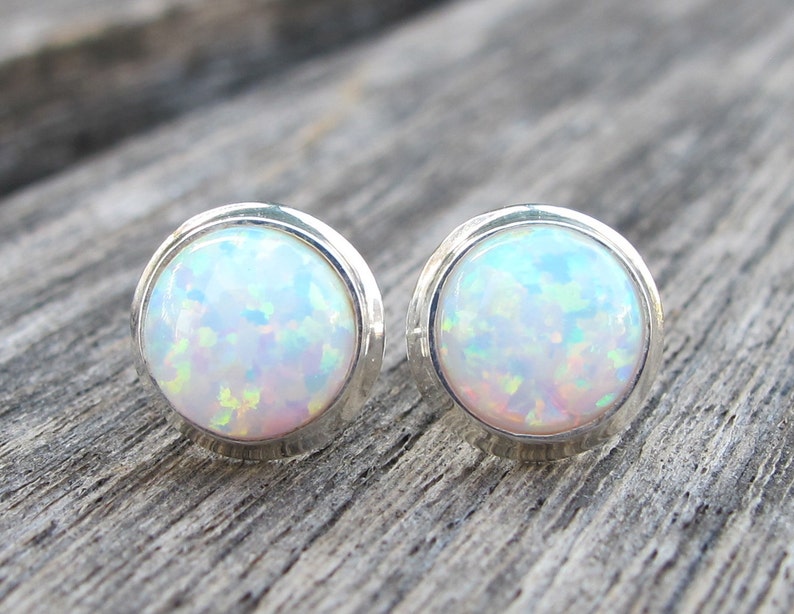 May include: A pair of round stud earrings featuring iridescent opal stones set in silver bezels. The opals display a mix of colors, including white, blue, and hints of pink and green. The earrings are displayed on a wooden surface.