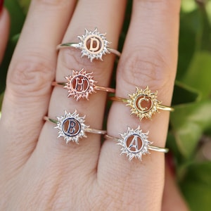 Puede incluir: Anillos personalizados con diseño de sol alrededor de una inicial. Los anillos son de plata, oro y oro rosa. Se ven las iniciales D, H, B, C y A. Joyería personalizada.