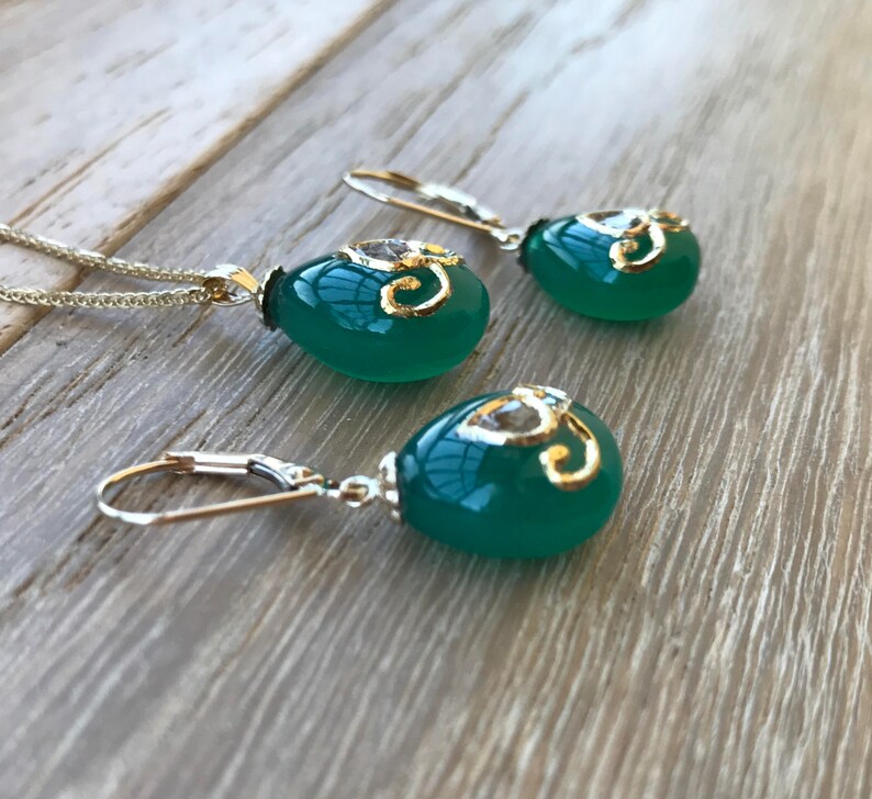 Op de afbeelding: Een ketting met een groene edelsteen hanger en bijpassende oorbellen met gouden accenten en een helder kristal. De hanger en oorbellen zijn traanvormig.
