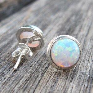 May include: A pair of round opal stud earrings. Each earring features a vibrant, iridescent opal stone set in a polished silver bezel. The opals display a mix of colors, including blue, green, and orange. The earrings are displayed on a wooden surface.