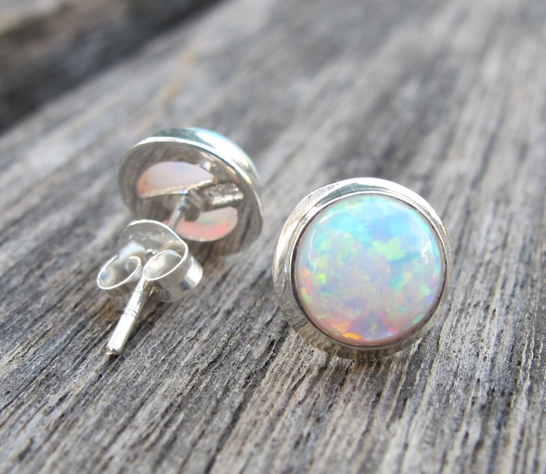 May include: A pair of round opal stud earrings. Each earring features a vibrant, iridescent opal stone set in a polished silver bezel. The opals display a mix of colors, including blue, green, and orange. The earrings are displayed on a wooden surface.