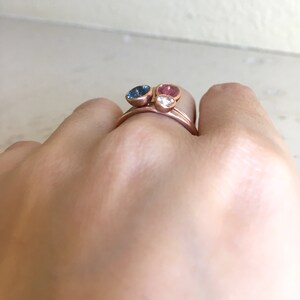 May include: Rose gold rings with three gemstone settings. The gemstones are a blue, pink, and clear stone. The rings are stacked on a finger.