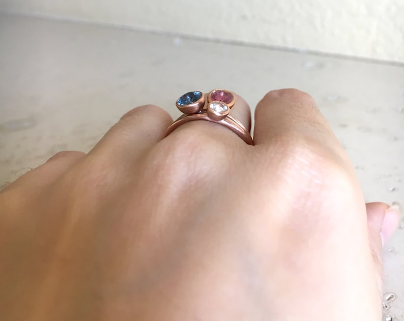 May include: Rose gold rings with three gemstone settings. The gemstones are a blue, pink, and clear stone. The rings are stacked on a finger.