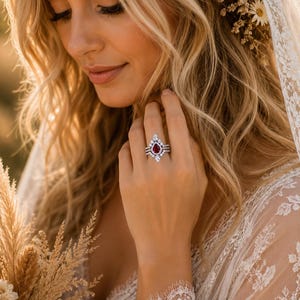 May include: A silver-toned ring set featuring a central pear-shaped red gemstone surrounded by a halo of small clear stones. The band is adorned with additional clear stones in a symmetrical arrangement, creating a layered, multi-stone appearance.
