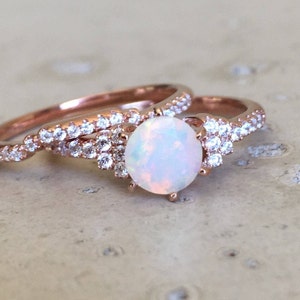Pode incluir: Um anel de noivado de ouro rosa com um grande opala branco oval e pedras transparentes menores. A banda também é adornada com pedras transparentes.