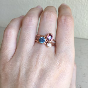 May include: A hand wearing four rose gold rings. Each ring features a different gemstone: a blue oval, a pink oval, and a white round stone. The rings are stacked on the finger.