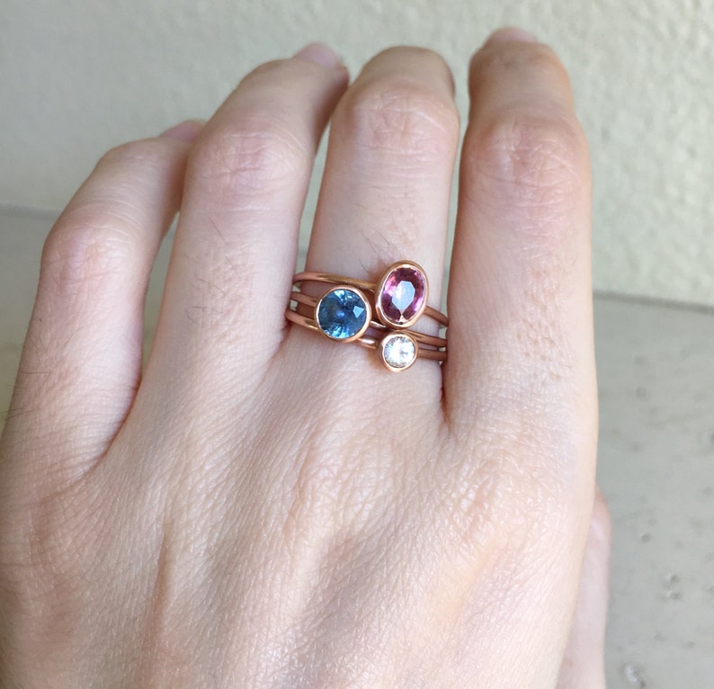 May include: A hand wearing four rose gold rings. Each ring features a different gemstone: a blue oval, a pink oval, and a white round stone. The rings are stacked on the finger.