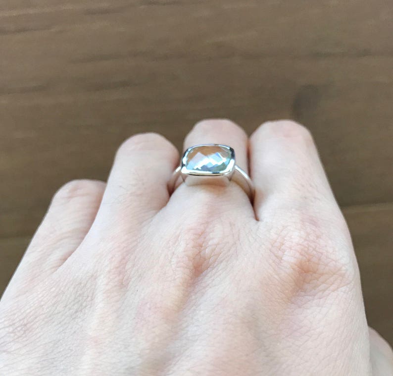 May include: A silver ring with a clear, faceted gemstone. The ring is on a person's finger.