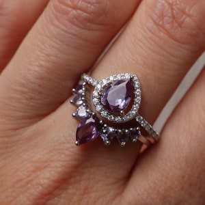 May include: A silver ring with a large pear-shaped amethyst gemstone surrounded by smaller diamonds. The ring is set on a band with a row of smaller amethyst gemstones.
