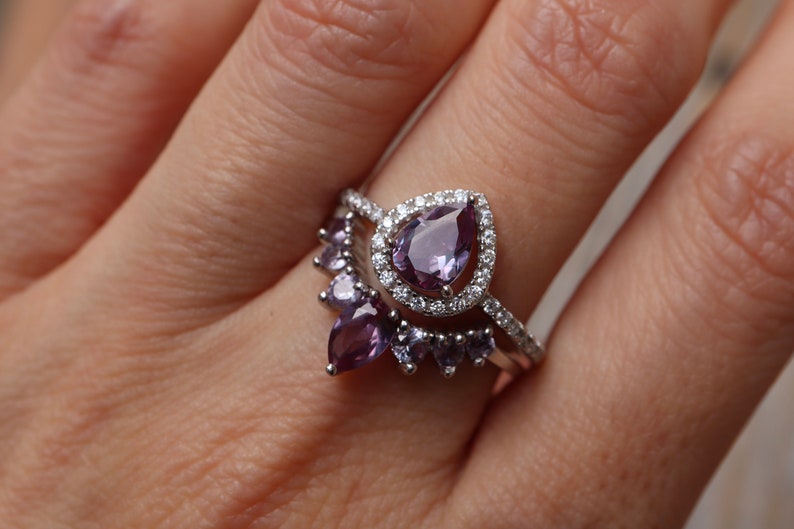 May include: A silver ring with a large pear-shaped amethyst gemstone surrounded by smaller diamonds. The ring is set on a band with a row of smaller amethyst gemstones.