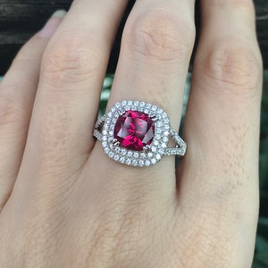 May include: A silver ring with a large, square-shaped, red gemstone surrounded by a double halo of smaller, clear gemstones.