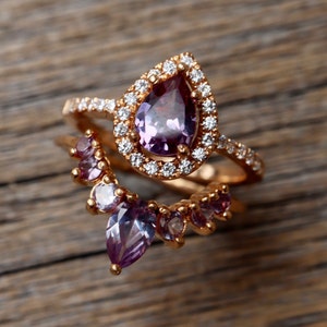 May include: Two gold rings with purple and clear gemstones. The larger ring features a pear-shaped purple gemstone surrounded by a halo of clear gemstones. The smaller ring has a curved design with multiple purple and clear gemstones.
