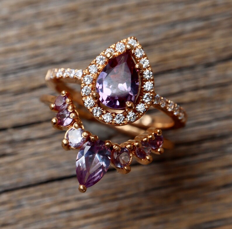 May include: Two gold rings with purple and clear gemstones. The larger ring features a pear-shaped purple gemstone surrounded by a halo of clear gemstones. The smaller ring has a curved design with multiple purple and clear gemstones.