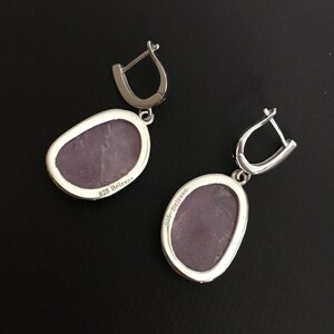 K&ouml;nnte beinhalten: Ein Paar silberne Ohrh&auml;nger mit einem violetten Stein. Die R&uuml;ckseite des Steins ist mit "925 Belenos" gekennzeichnet.