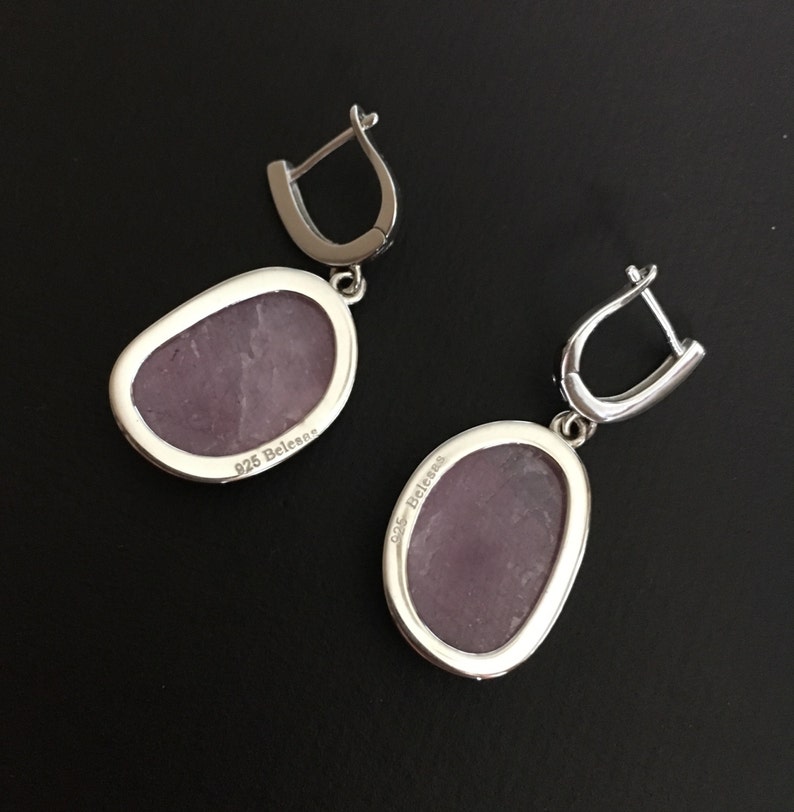 K&ouml;nnte beinhalten: Ein Paar silberne Ohrh&auml;nger mit einem violetten Stein. Die R&uuml;ckseite des Steins ist mit "925 Belenos" gekennzeichnet.