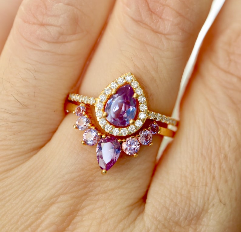 May include: A gold ring set with a pear-shaped purple gemstone surrounded by smaller clear gemstones. A second band with smaller purple gemstones is worn below the main ring.