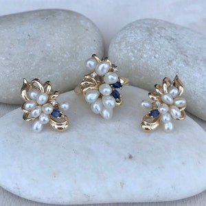 May include: A set of gold-tone jewelry featuring a ring and earrings. The pieces are adorned with a cluster of white pearls and blue gemstones.