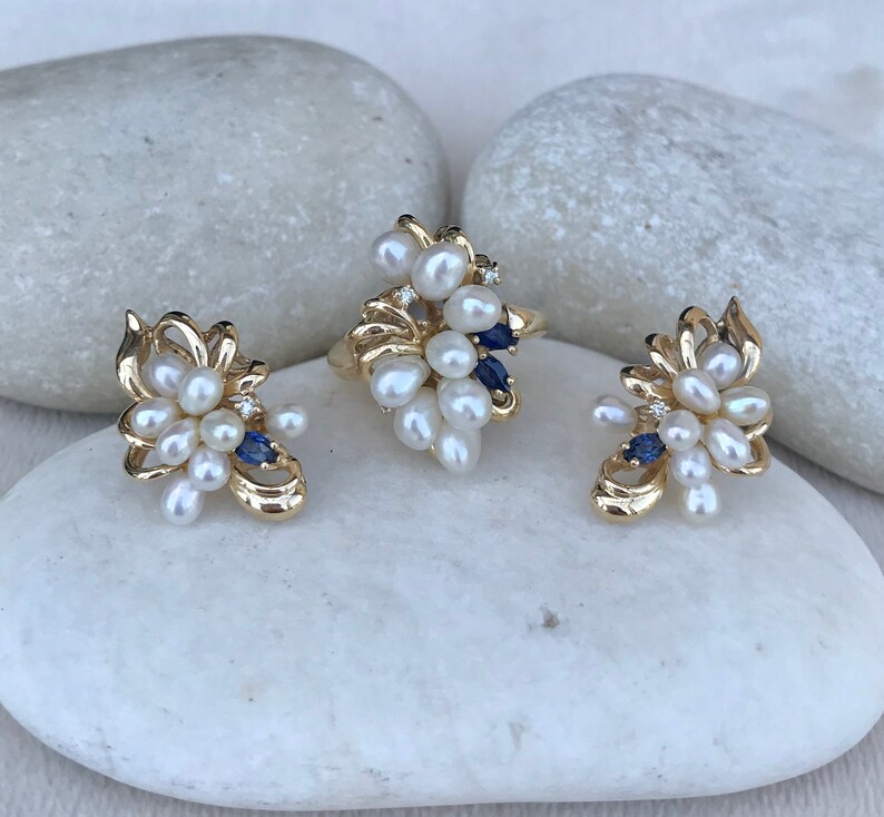 May include: A set of gold-tone jewelry featuring a ring and earrings. The pieces are adorned with a cluster of white pearls and blue gemstones.