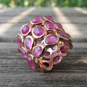 Pode incluir: Um anel de ouro com várias filas de pedras preciosas ovais cor-de-rosa. O anel está colocado sobre uma superfície de madeira.