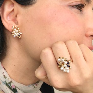May include: A gold ring and earrings set with a cluster of white pearls and blue gemstones. The ring is on a finger and the earrings are in the earlobes.