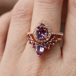 May include: A rose gold ring with a pear-shaped amethyst gemstone surrounded by a halo of smaller diamonds. The ring is set with a band of smaller amethysts.
