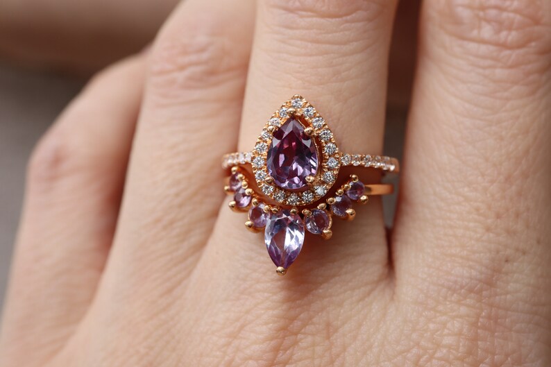 May include: A rose gold ring with a pear-shaped amethyst gemstone surrounded by a halo of smaller diamonds. The ring is set with a band of smaller amethysts.