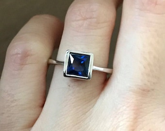 May include: A silver ring with a square-shaped blue gemstone set in a silver bezel.