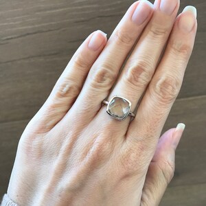 May include: A silver ring with a clear, square-shaped gemstone. The ring is on a person's finger.