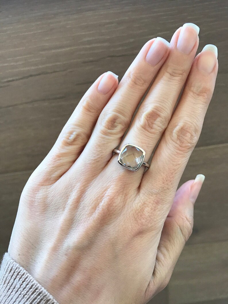 May include: A silver ring with a clear, square-shaped gemstone. The ring is on a person's finger.