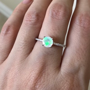 Pode incluir: Um anel de prata com uma pedra de opala redonda e iridescente e uma banda de pequenas pedras transparentes.