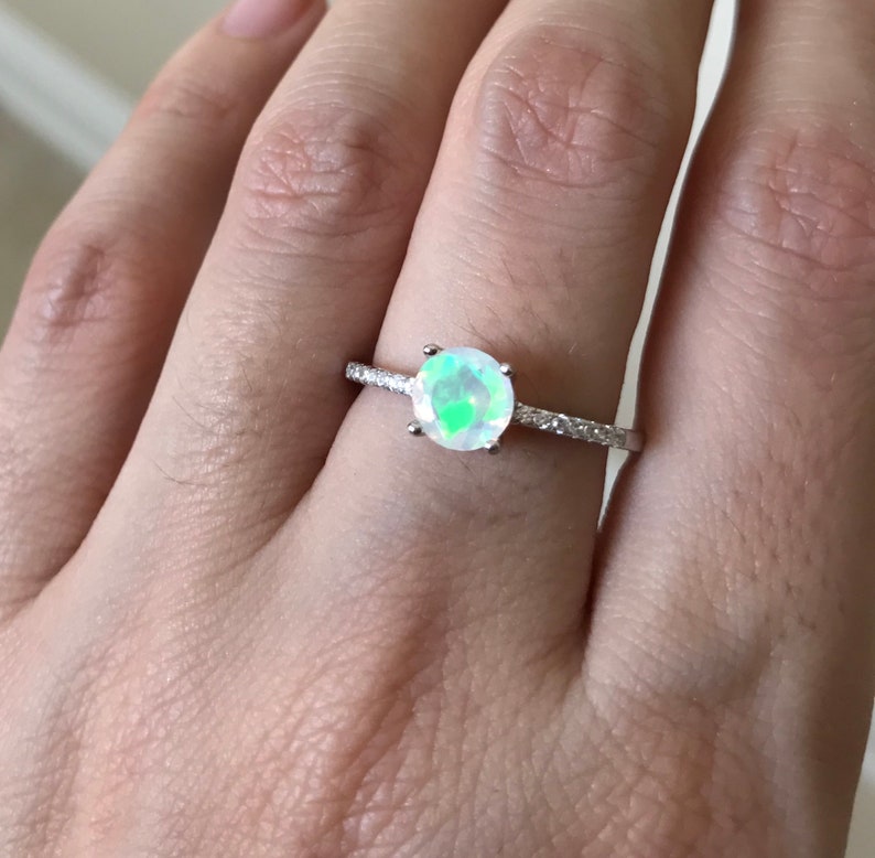 Pode incluir: Um anel de prata com uma pedra de opala redonda e iridescente e uma banda de pequenas pedras transparentes.
