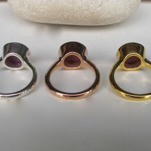 Pu&ograve; includere: Tre anelli con una pietra preziosa viola incastonata in una montatura d'argento, oro rosa e oro. Gli anelli sono mostrati dal retro, mostrando le bande metalliche lisce.