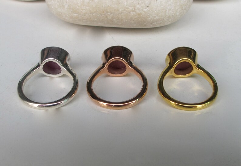 Pu&ograve; includere: Tre anelli con una pietra preziosa viola incastonata in una montatura d'argento, oro rosa e oro. Gli anelli sono mostrati dal retro, mostrando le bande metalliche lisce.
