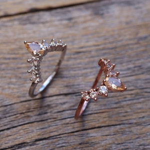 May include: Two delicate rings with a V-shaped design featuring pear-shaped gemstones and smaller round stones. The ring on the left is silver, and the ring on the right is rose gold.