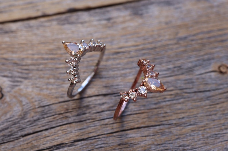 May include: Two delicate rings with a V-shaped design featuring pear-shaped gemstones and smaller round stones. The ring on the left is silver, and the ring on the right is rose gold.