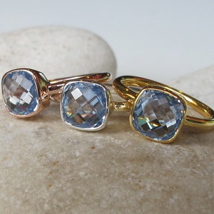 May include: Three rings with square-shaped blue gemstones set in gold, rose gold, and silver settings.