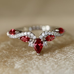 May include: A silver ring with a floral design, featuring a large, pear-shaped red gemstone at the center, surrounded by smaller red gems and clear, sparkling stones. The ring is set on a soft, beige surface.