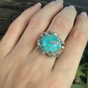 Op de afbeelding: Een zilveren ring met een grote turquoise steen omringd door kleinere blauwe stenen.