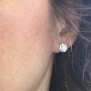 May include: A close-up of a person's ear with a small, round, white stud earring. The earring is set in a silver or light-colored metal. The person's skin is fair, and their hair is dark and wavy.