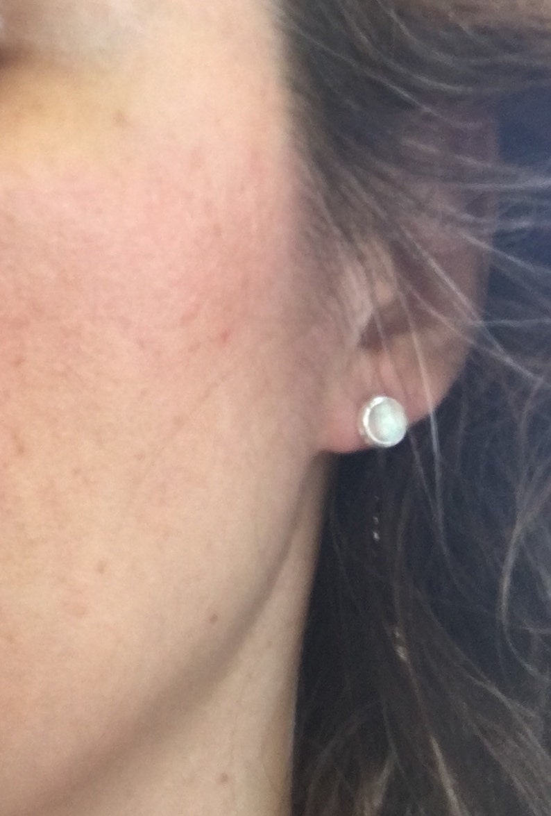 May include: A close-up of a person's ear with a small, round, white stud earring. The earring is set in a silver or light-colored metal. The person's skin is fair, and their hair is dark and wavy.