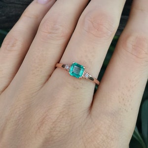 Op de afbeelding: Een ring van ros&eacute;goud met een vierkante smaragdgroene edelsteen en twee kleinere ronde diamanten aan weerszijden.