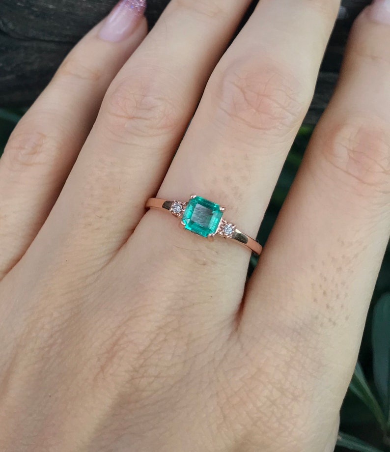 Op de afbeelding: Een ring van ros&eacute;goud met een vierkante smaragdgroene edelsteen en twee kleinere ronde diamanten aan weerszijden.
